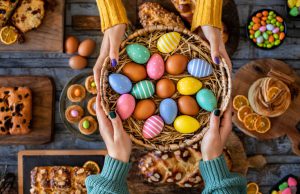 mãos femininas segurando cestas com diversos ovos coloridos de páscoa e ao fundo diversos doces
